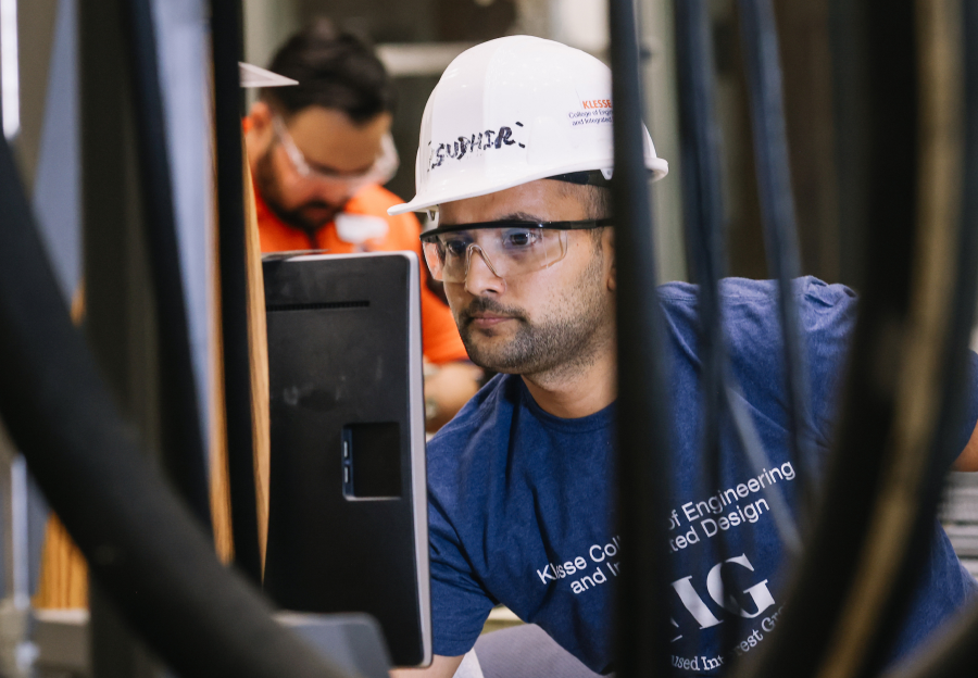 A male engineering student wearing a white hard hat, working on a computer