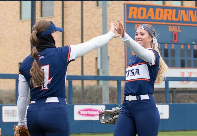 A young woman in a softball uniform high fives another person.