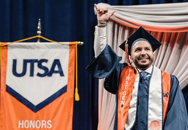 A student on the graduation stage with a 'Birds Up' hand sign, next to a 'UTSA' orange banner behind him.