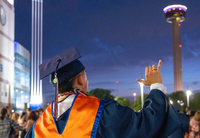 A male UT San Antonio graduate stands outside the Alamodome while looking up at the Tower of the Americas and doing the 'Birds' up hand sign